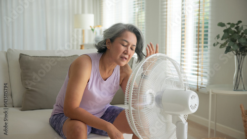 Asia adult people middle age old woman sit at sofa home feel hot flash fever use fan waving hand blowing wind face elder health care summer heat aging issue. Sad pain sweat tired stroke suffer stress.