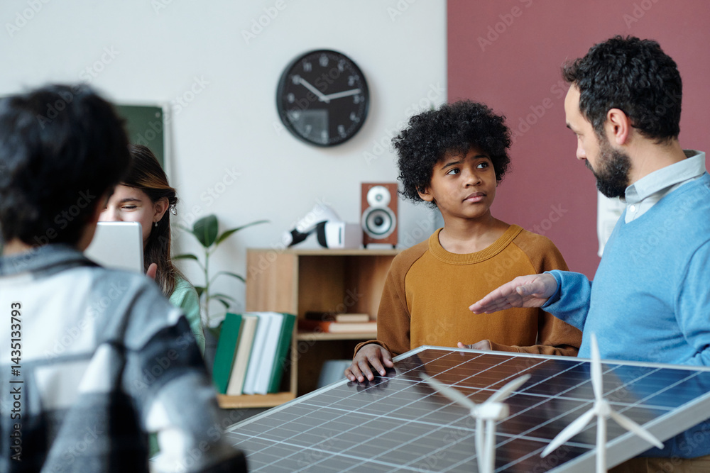 Fototapeta premium Group of diverse individuals discussing renewable energy models in classroom setting with teacher showing solar panel and various educational materials visible in background