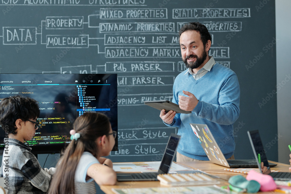 custom made wallpaper toronto digitalTeacher in blue sweater teaching children coding concepts using tablets and laptops in modern classroom setting with chalkboard background showcasing programming terms