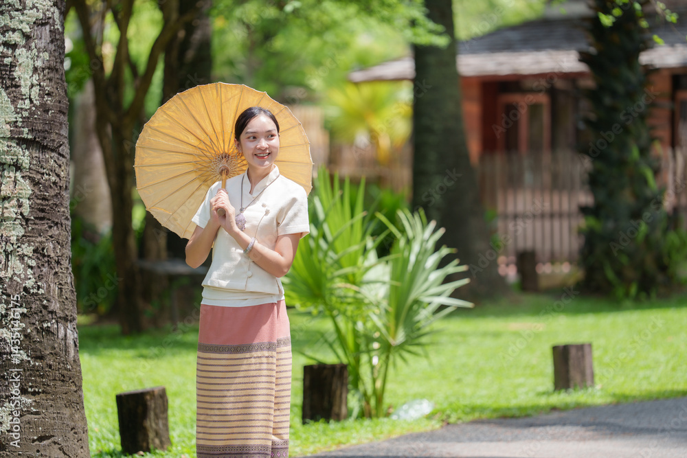 Naklejka premium Beautiful thai woman in traditional clothing holding a parasol and smiling in a serene temple garden, showcasing the rich culture and peaceful atmosphere of thailand