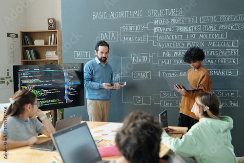 Instructor explaining algorithmic structures to attentive students using chalk and smartboard in classroom. Group of diverse students engaged in learning coding concepts
