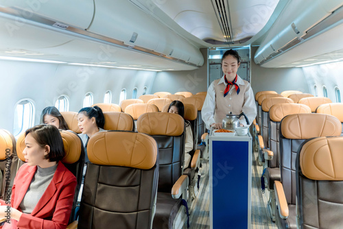 Flight attendant pushing food service cart in airplane aisle, providing in-flight meal and beverage service to passengers seated in economy class cabin