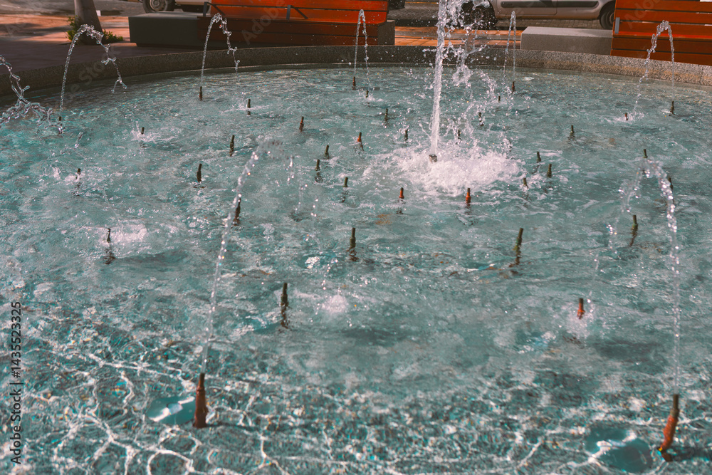 Circular fountain with multiple jets sprays water upward, creating splashes and ripples in shallow pool. Red benches and paved walkway surround fountain, adding vibrant contrast to water blue tones