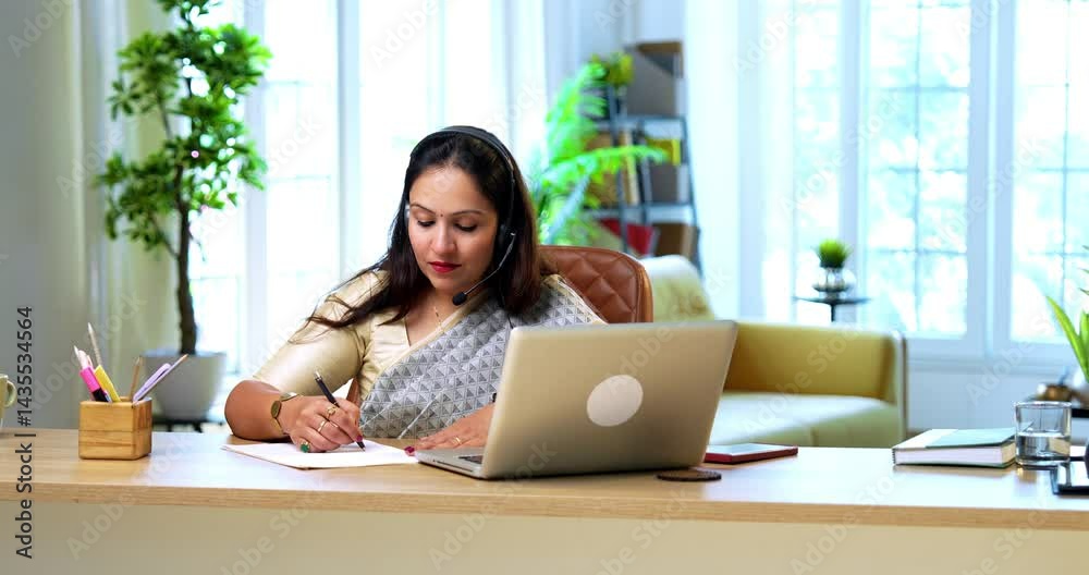 Speaking in video call with headphones and taking notes on paper, Indian young businesswoman or female corporate lady in saree attending online meeting on laptop at cozy modern office desk Speaking in video call with headphones and taking notes on paper, Indian young businesswoman or female corporate lady in saree attending online meeting on laptop at cozy modern office desk