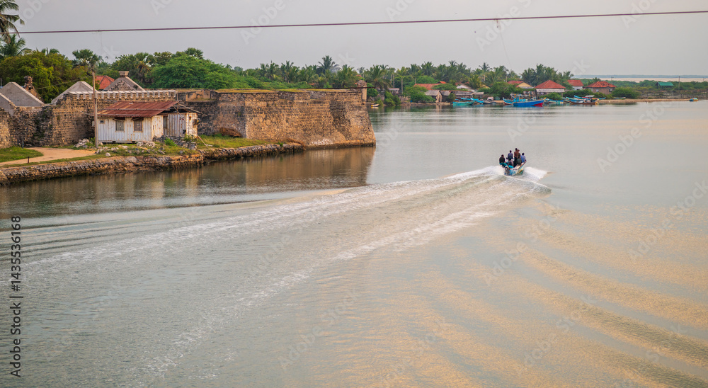 Obraz premium A small fisherman boat speeds across the calm waters near the historic Mannar Dutch Fort in Sri Lanka, leaving a rippling trail behind under a golden sunset sky