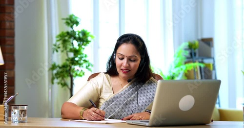 Writing notes in diary while working on laptop, young Indian businesswoman in saree focused on multitasking office work, sitting at cozy desk in corporate workplace using modern technology