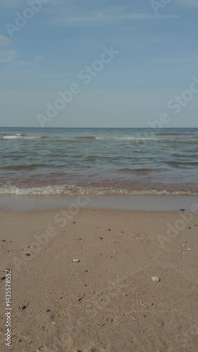 Peaceful seaside landscape of Baltic sea coast with calm turquoise waves rolling on empty sandy beach under clear blue sky at summer day. With no people vertical seascape natural background.