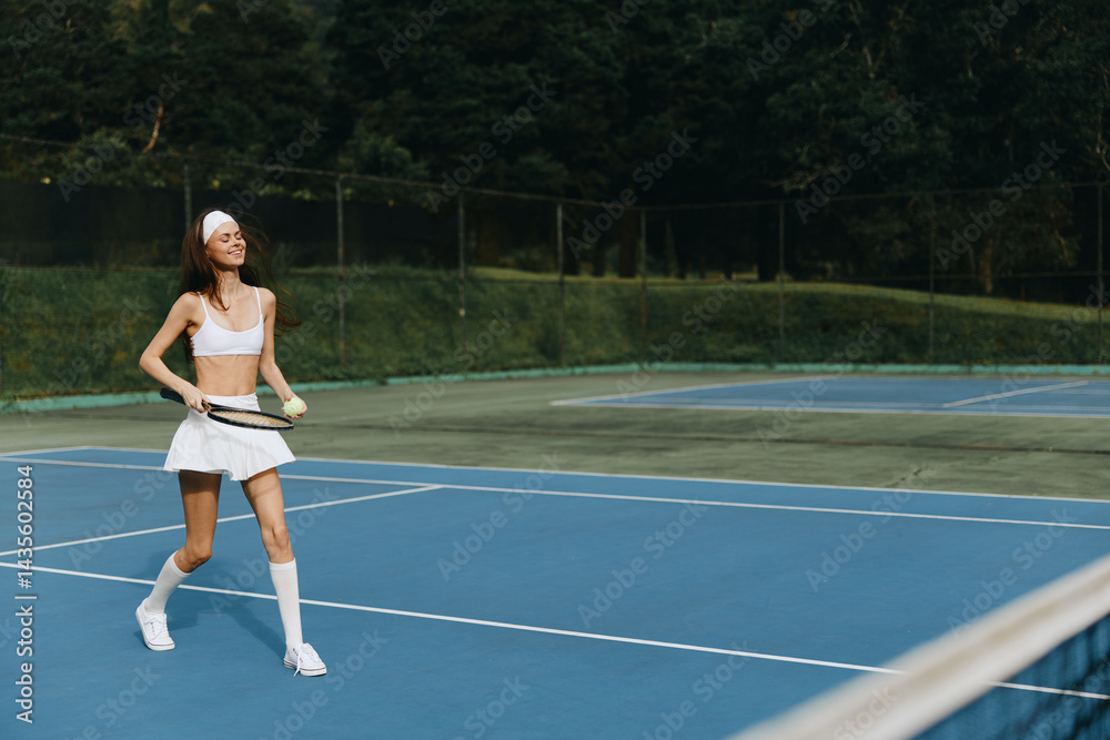 Obraz premium Young athletic woman playing tennis outdoors in a stylish white sport outfit, showcasing joy and energy on a blue court surrounded by nature