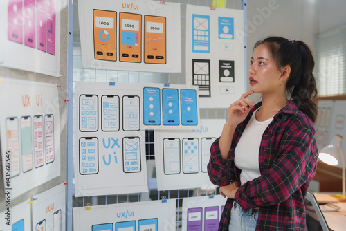 Uxui designer examines mobile app prototypes on a wall, focusing on layout and functionality. The workspace is filled with sketches and design plans, highlighting the creative process photo