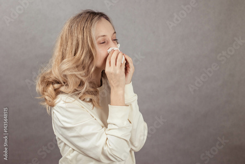 portrait of unhealthy cute blonde female in white top with napkin blowing nose, copy space, allergy concept