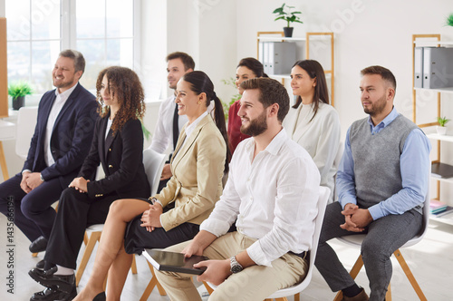 Group of smiling young business people sitting in a row on chairs and listening to colleague or their leadership at working place during business training or conference in meeting room.