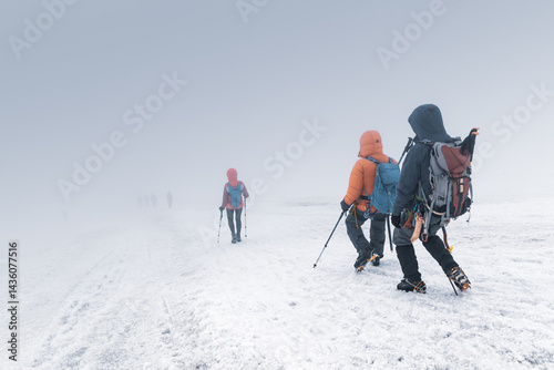 Climbers with backpacks, trekking poles, crampons walk down snow covered mountain slope in dense fog