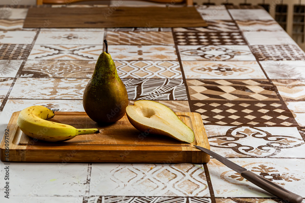 Rustic fruit still life: Pear, banana, cut pear on board with knife. Clear light on patterned table.