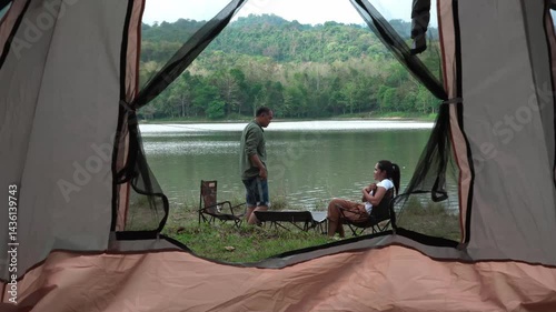 man and a woman are setting up a tent beside a peaceful lake surrounded by lush green forest and hills. The scene captures the essence of outdoor adventure and camping in a serene, natural environment