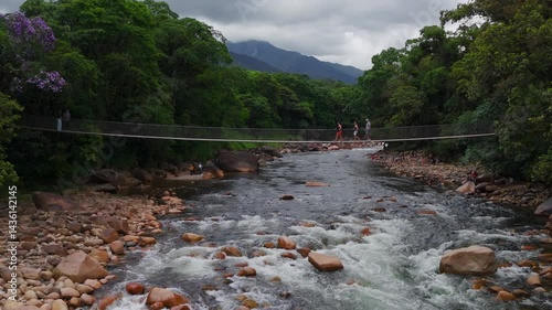 Rustic wooden cable bridge spans a tranquil river nestled in green mountains. Lush nature surrounds this peaceful crossing