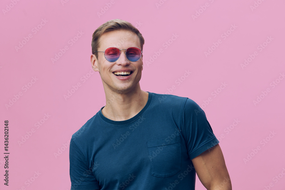 Smiling young man wearing colorful sunglasses blue t-shirt looking away happy casual lifestyle concept pink background Smiling young man wearing colorful sunglasses blue t-shirt looking away happy casual lifestyle concept pink background