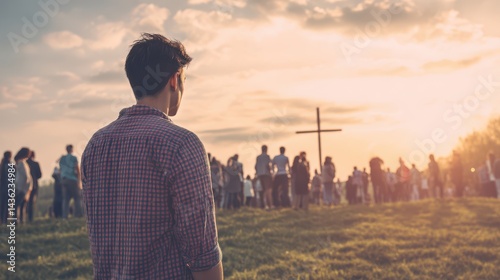 Young man looking at a crowd of people praying in front of a cross on a meadow with sunset light