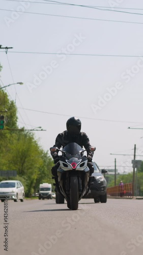 Man driver in black leather jacket and helmet rides powerful bike along large road with cars in modern city on sunny day front view slow motion