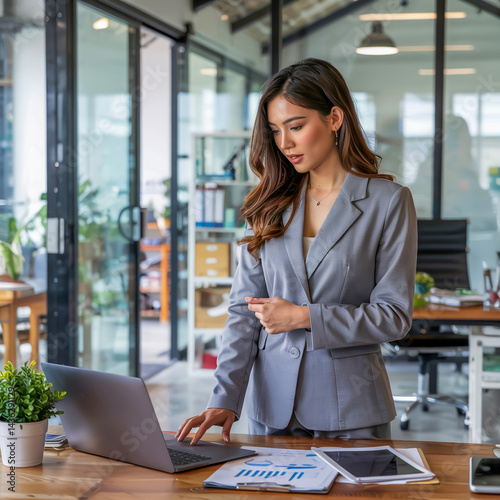 Young woman in a chic gray pantsuit reviewing documents on a laptop in a minimalist office, floor-to-ceiling windows, professional mood