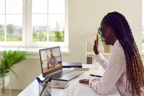 Obraz na płótnie Diverse business woman and man having online work meeting via video call. Happy African American woman financial accountant sitting at her office desk, smiling and waving hello at laptop computer w salonie