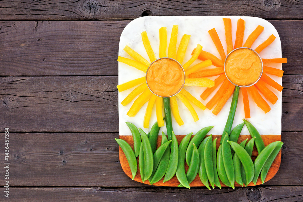 Fun summer theme vegetable snack board against a dark wood background. Flower arranged veggies with hummus. Above view.