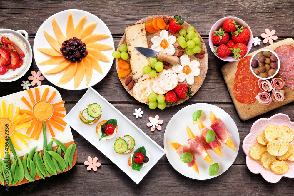 Summer garden party food table scene. Assortment of fun flower theme charcuterie items, cheese, fruit, vegetables and finger snacks. Top down view on a dark wood background.