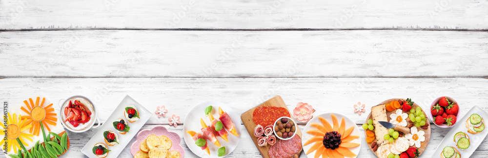 Summer garden party food bottom border. Assortment of fun flower theme charcuterie items, cheese, fruit, vegetables and finger snacks. Top down view on a white wood banner background.