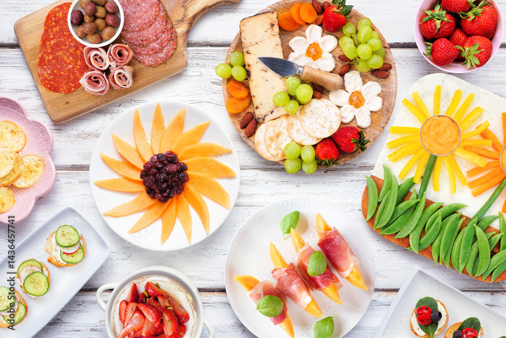 Summer garden party food table scene. Variety of fun flower theme charcuterie items, cheese, fruit, vegetables and finger snacks. Overhead view on a white wood background.