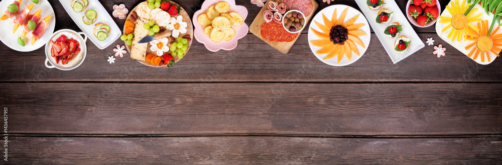 Summer garden party food top border. Assortment of fun flower theme charcuterie items, cheese, fruit, vegetables and finger snacks. Above view on a dark wood banner background.