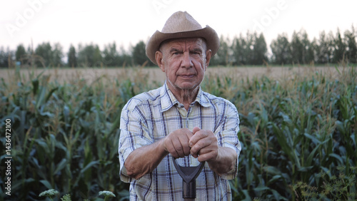 Portrait of elderly old farmer man with deep wrinkles on his skin and hat on head. He is standing outdoors at cornfield of rural field holding shovel in his hands.