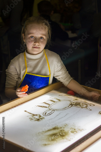 Beautiful little girl draws with sand on a light square, art therapy, sand animation, creative leisure for children, selective focus	