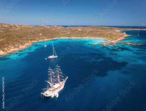 Aerial view of sail boat and luxury yachts on blue sea at sunset in summer. Sardinia, Italy. Tropical seascape with speed boats, yachts, sea bay, mountain, clear azure water, sky. Top view of ocean