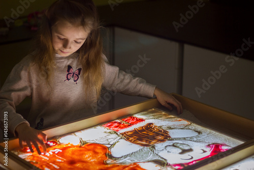 Beautiful little girl draws with sand on a light square, art therapy, sand animation, creative leisure for children, selective focus