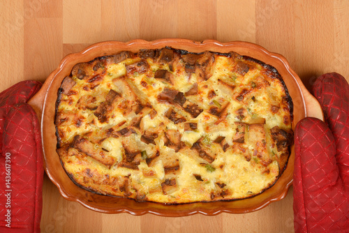 Obraz na płótnie A freshly baked strata (layered casserole dish made from a mixture which mainly consists of bread, eggs and cheese) is being held with oven mitts on a wooden surface w salonie