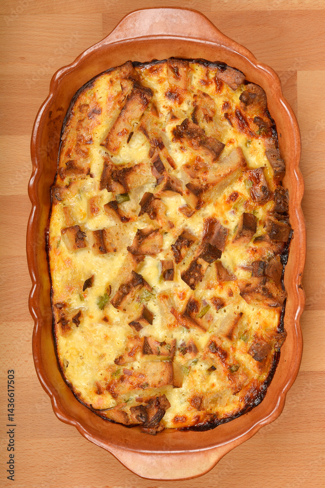 A baked strata (layered casserole dish made from a mixture which mainly consists of bread, eggs and cheese) in a rustic dish sits on a wooden table, ready to be served