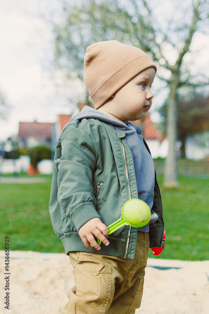 A boy of 2 years old in a spring jacket and hat plays with sand in a sandbox. A boy plays on a playground with a shovel in the sand. A child's walk in the fresh air