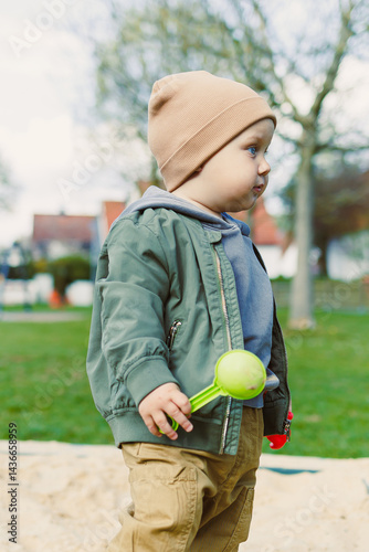 Obraz na płótnie A boy of 2 years old in a spring jacket and hat plays with sand in a sandbox. A boy plays on a playground with a shovel in the sand. A child's walk in the fresh air w salonie