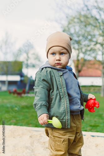 A boy of 2 years old in a spring jacket and hat plays with sand in a sandbox. A boy plays on a playground with a shovel in the sand. A child's walk in the fresh air