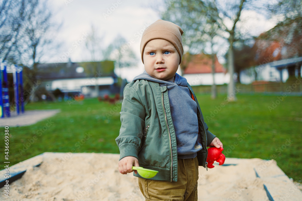 A boy of 2 years old in a spring jacket and hat plays with sand in a sandbox. A boy plays on a playground with a shovel in the sand. A child's walk in the fresh air
