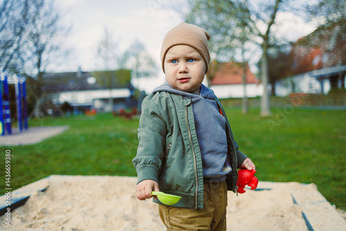 A boy of 2 years old in a spring jacket and hat plays with sand in a sandbox. A boy plays on a playground with a shovel in the sand. A child's walk in the fresh air