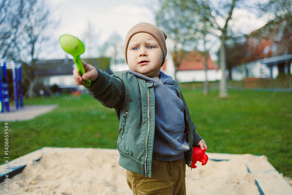 A boy of 2 years old in a spring jacket and hat plays with sand in a sandbox. A boy plays on a playground with a shovel in the sand. A child's walk in the fresh air