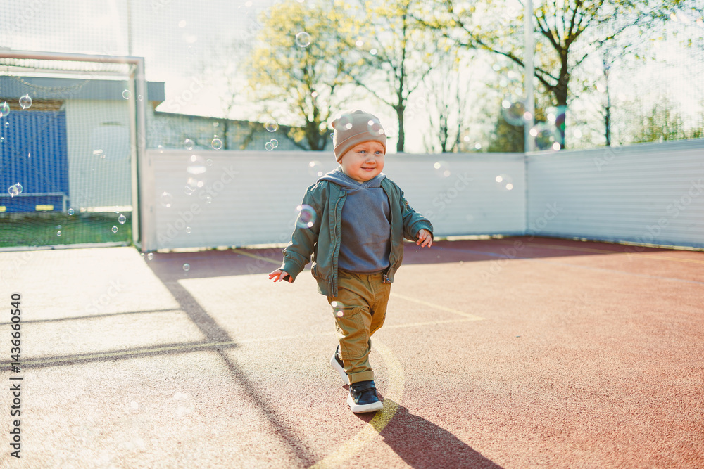 Toddler boy running outside with soap bubbles. Child on a warm spring day for a walk in a jacket and hat.