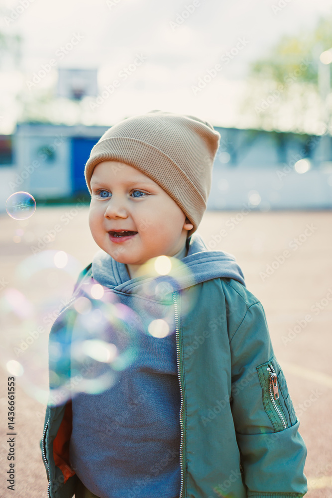 Toddler boy running outside with soap bubbles. Child on a warm spring day for a walk in a jacket and hat.