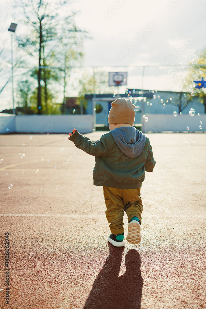 Cheerful 2 year old boy playing outside in summer in a green jacket with soap bubbles. Toddler boy running outside and playing with bubbles.
