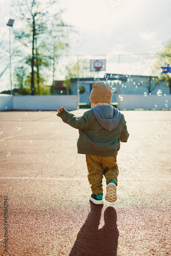 Obraz na płótnie Cheerful 2 year old boy playing outside in summer in a green jacket with soap bubbles. Toddler boy running outside and playing with bubbles. w salonie