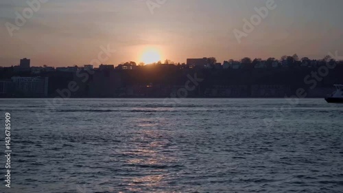 A breathtaking sunset view of a city skyline silhouetted against a vibrant sky, with warm hues of orange and pink blending into cool blue over a calm body of water, creating a serene urban landscape.