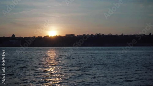 A breathtaking sunset view of a city skyline silhouetted against a vibrant sky, with warm hues of orange and pink blending into cool blue over a calm body of water, creating a serene urban landscape.