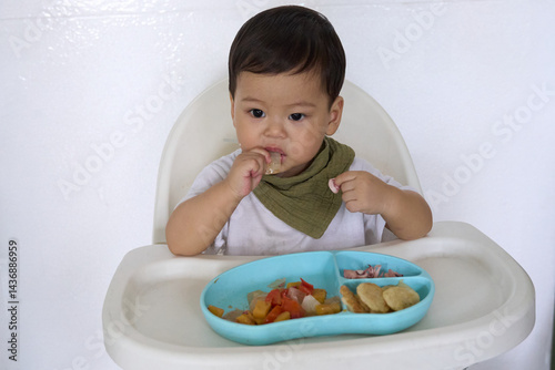 15 month old baby is sitting on high chair eating with fingers