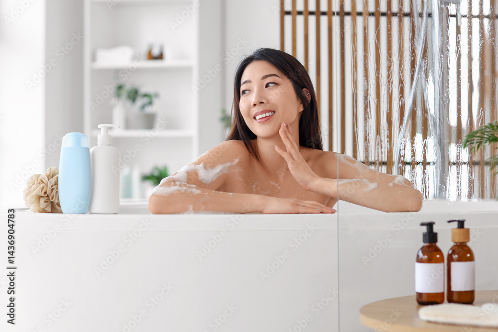 Pretty young Asian woman with bottles of shower gel and loofah taking bath in bathroom