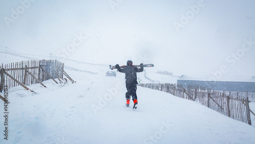 A skier carrying his skis walks through heavy blizzard conditions at Glencoe Mountain Resort in the Scottish Highlands. Thick snow falls heavily around him, coating his winter gear